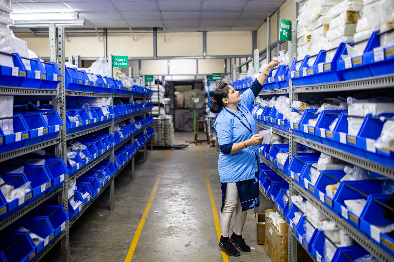 Warehouse worker in uniform picking medical stock from labelled blue bins in a pharmaceutical distribution aisle