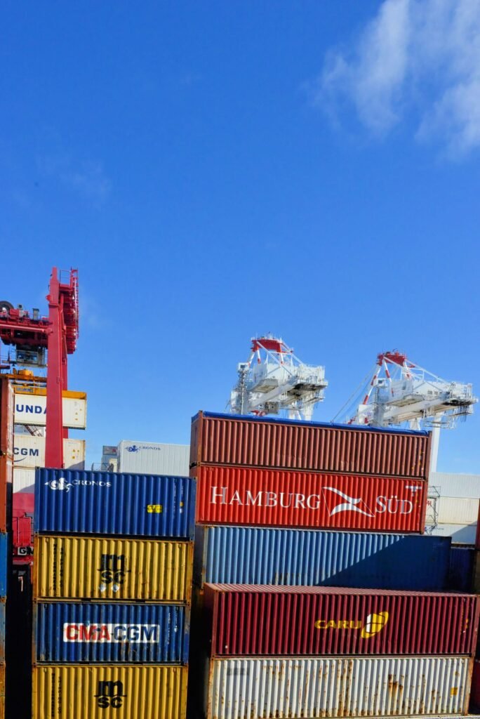 Stacked shipping containers from several carriers below dockside gantry cranes at a container port under a clear blue sky